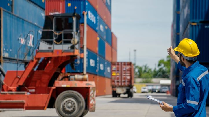 Port worker overseeing intermodal container loading with truck and reach stacker in the background
