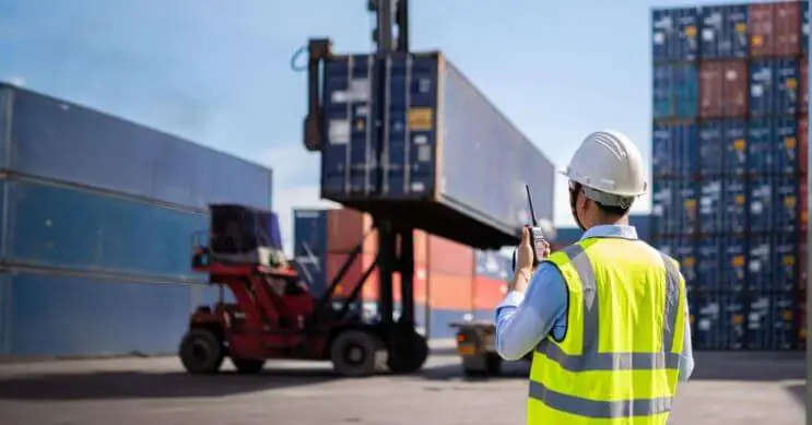 Um trabalhador portuário com capacete e colete de alta visibilidade segura um walkie-talkie enquanto monitoriza o processo de carregamento de contentores num porto movimentado. Em segundo plano, um empilhador levanta um contentor, o que garante o manuseamento adequado da carga e a eficiência do transporte.
