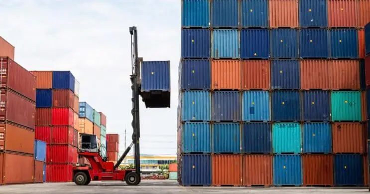 Um reach stacker a elevar um contentor carregado para uma pilha num terminal portuário movimentado. O manuseamento eficiente de contentores com reach stackers ajuda a otimizar as operações de carga, o que garante um empilhamento seguro e equilibrado dos contentores, enquanto melhora a eficiência do porto.