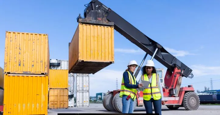 Two logistics workers discuss loading meter planning as a container is lifted by a reach stacker at a freight yard