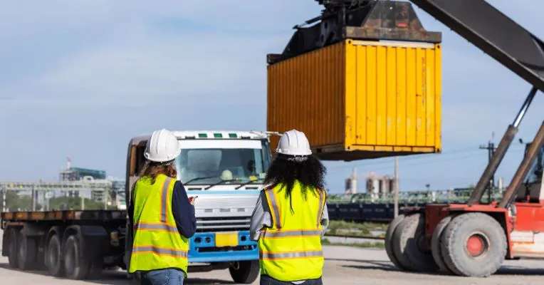 Freight load board - container handling in logistics Workers in safety vests and helmets supervising the loading of an intermodal container onto a truck, representing freight load board operations