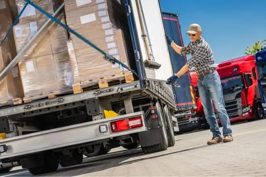 A worker in a plaid shirt, jeans, and a cap is securing cargo on a pallet in the back of a semi-truck. Other semi-trucks are parked nearby under a clear sky.