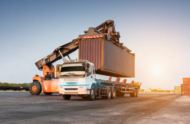 Truck being loaded with a shipping container at a freight yard, illustrating heavy cargo handling and the importance of accurate load weight calculation