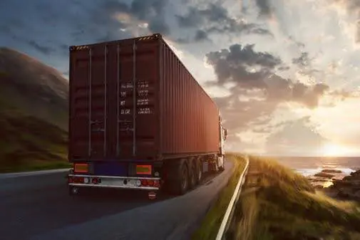 A large red shipping container on a truck drives along a coastal highway during sunset with the sunbeams shining through the clouds.