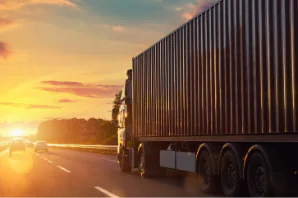 A large red shipping container truck drives along a highway during sunset