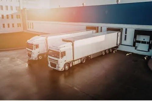 Two white semi-trucks are parked at loading docks outside a warehouse. The sun is setting, casting a warm glow over the scene.
