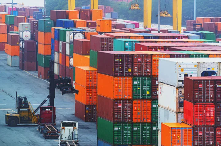 Stacked shipping containers in multiple colors at a container terminal, with a forklift operating between the rows.